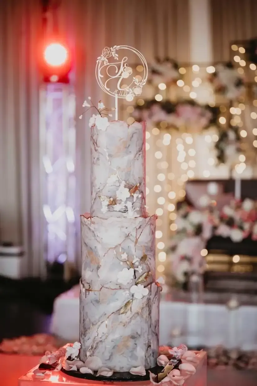 Tall marble-effect wedding cake with a gold circular “S&C”-style monogram topper, white florals and gold-leaf accents, at a reception with string lights