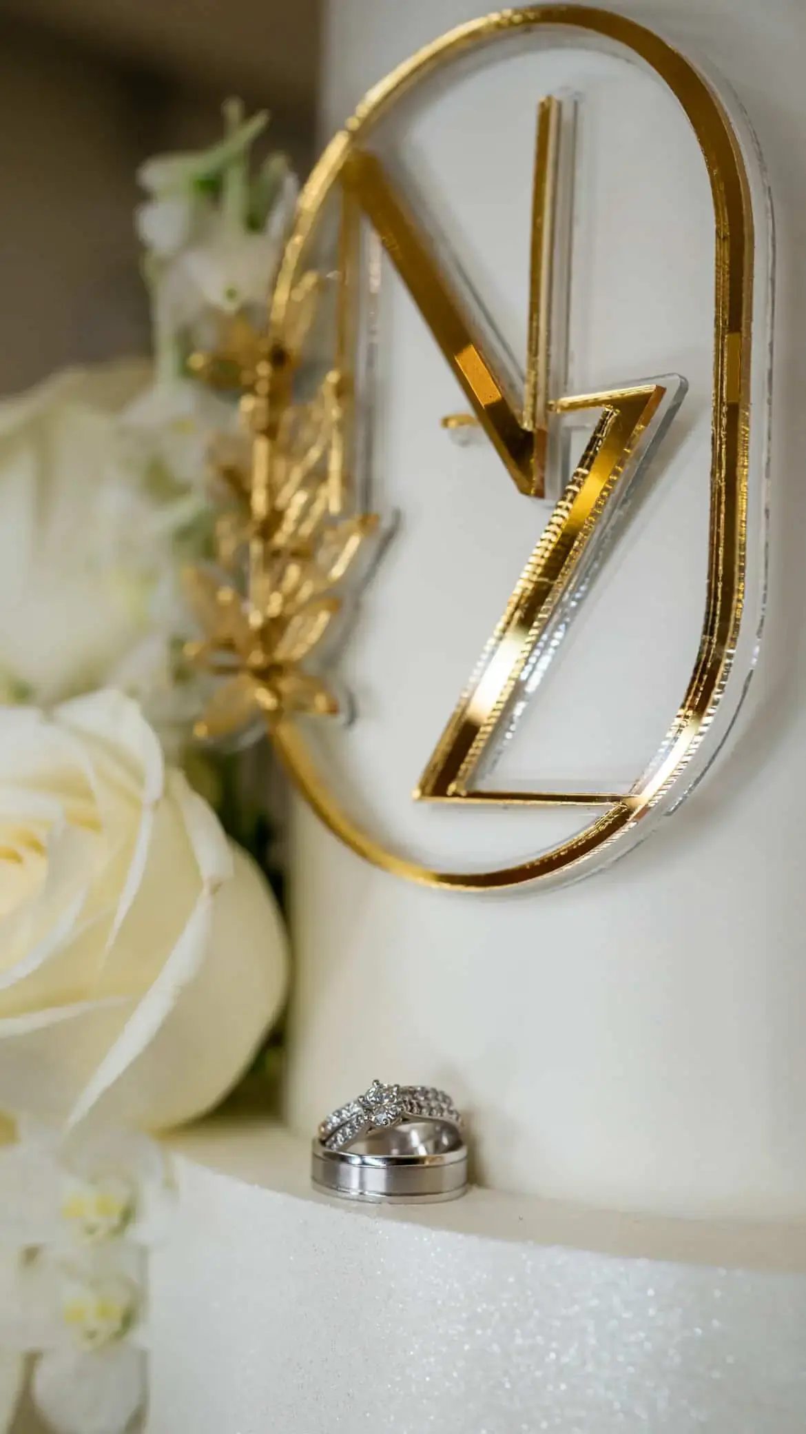 Close-up detail of a large gold monogram cake topper and a pair of wedding rings resting against a white iced tier, with white roses in the background