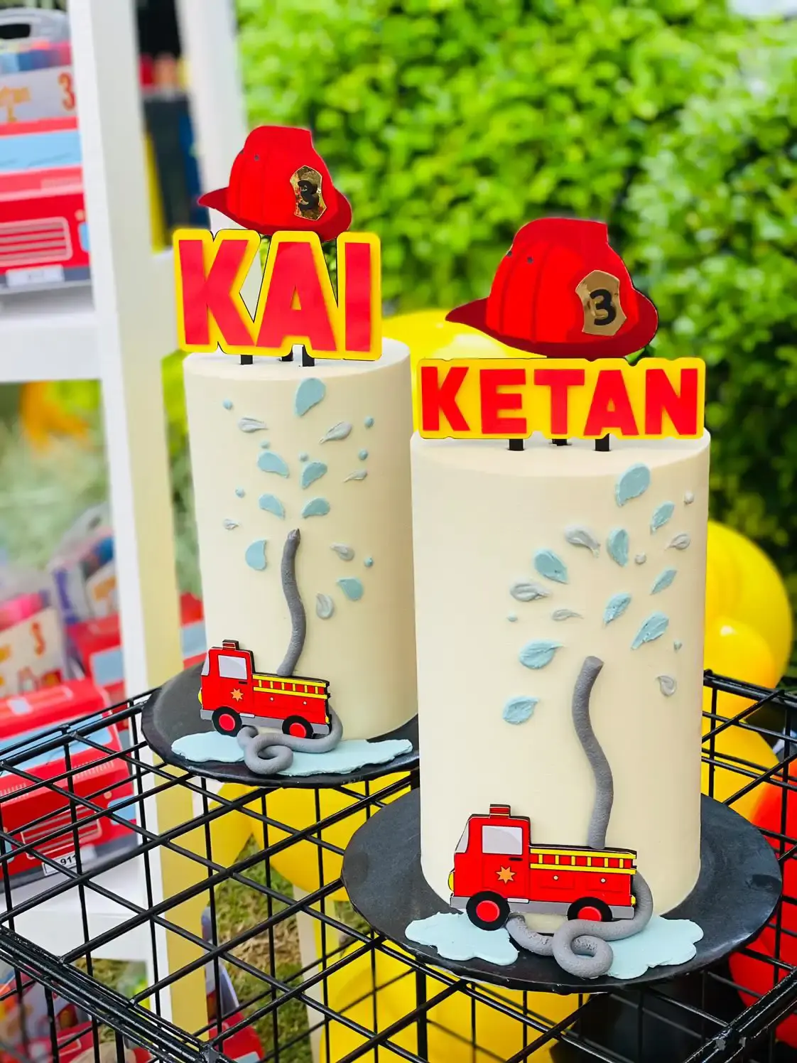 Two matching fire-truck themed birthday cakes with fireman-helmet toppers reading “Kai” and “Ketan” and piped water-splash icing