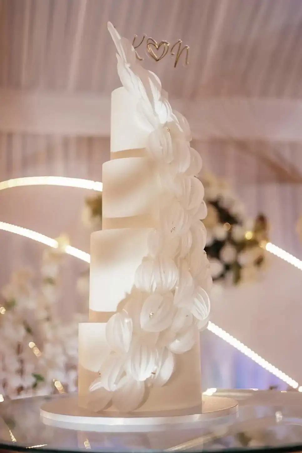 Tall white tiered wedding cake with a sculpted feather-texture cascade down one side and a heart-monogram topper, photographed at a string-lit reception