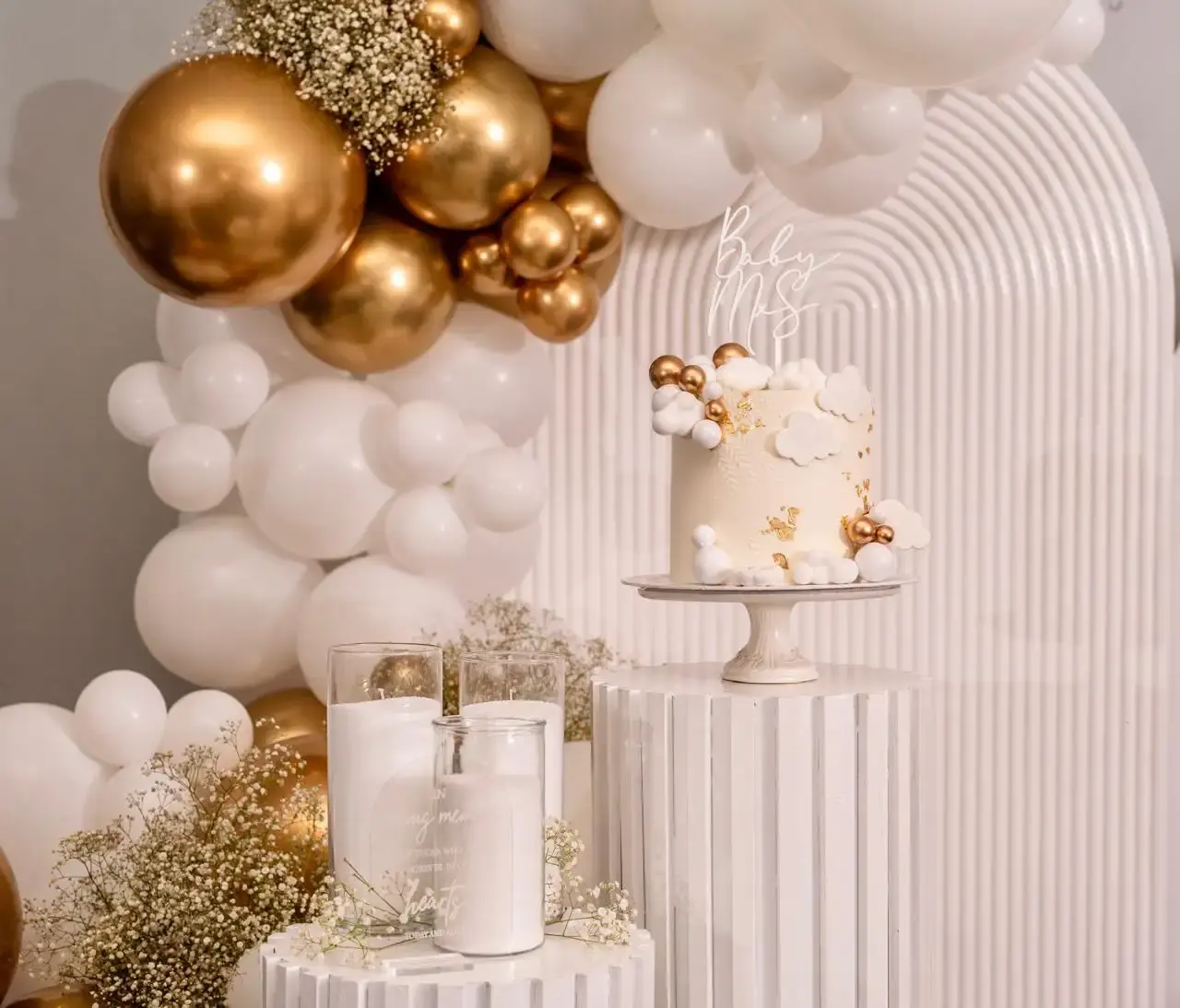 Wide shot of a cloud and gold-sphere “Baby Mrs” cake on a white fluted pedestal with candles in foreground