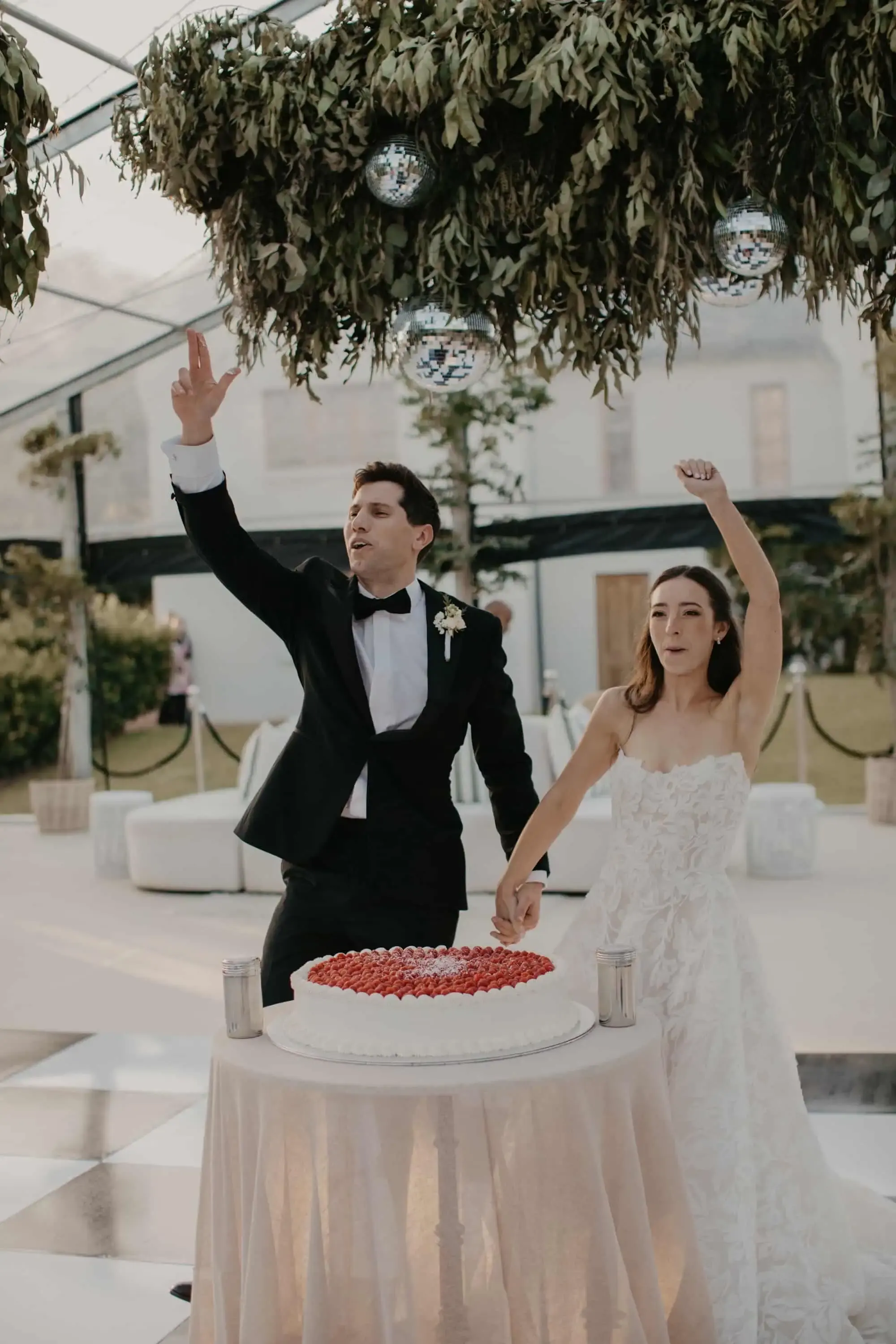 Bride and groom raising their arms in celebration beside a strawberry-topped cake on an outdoor dance floor, under a hanging greenery-and-disco-ball installation