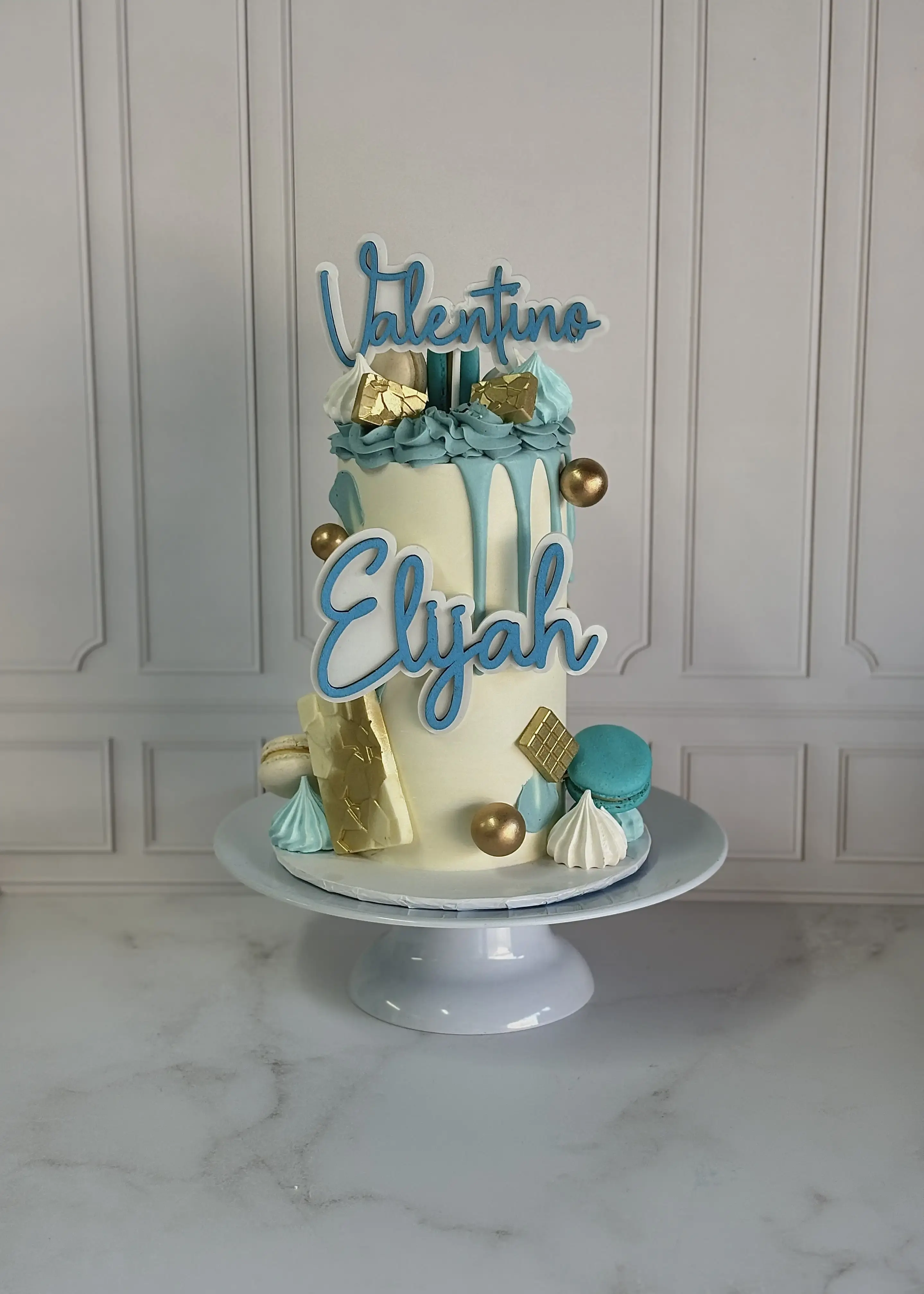 Cream cake with teal drip icing, macarons and meringue kisses, two names piped on acrylic toppers