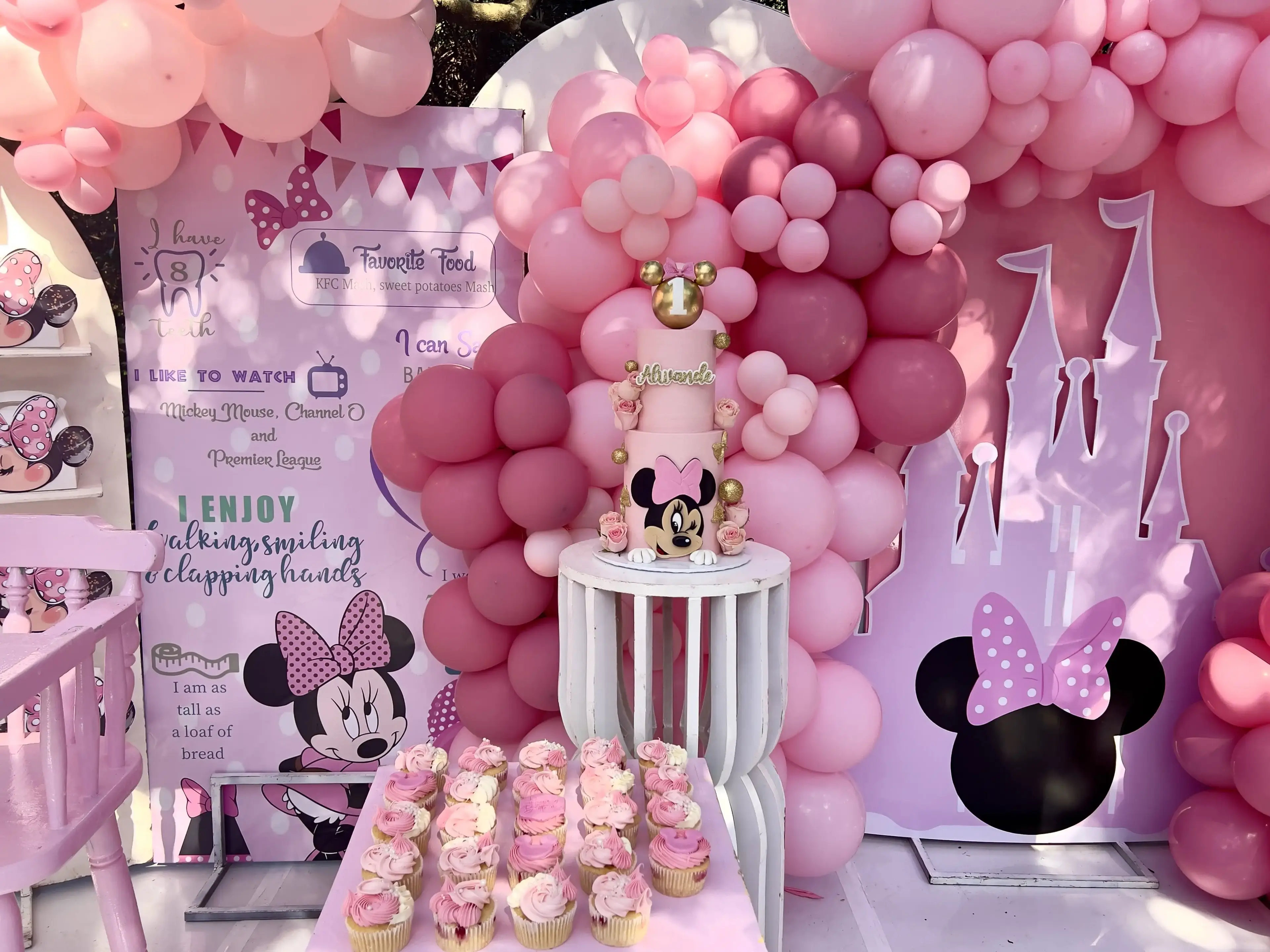 Pink two-tier Minnie Mouse first birthday cake with photo topper against a balloon garland