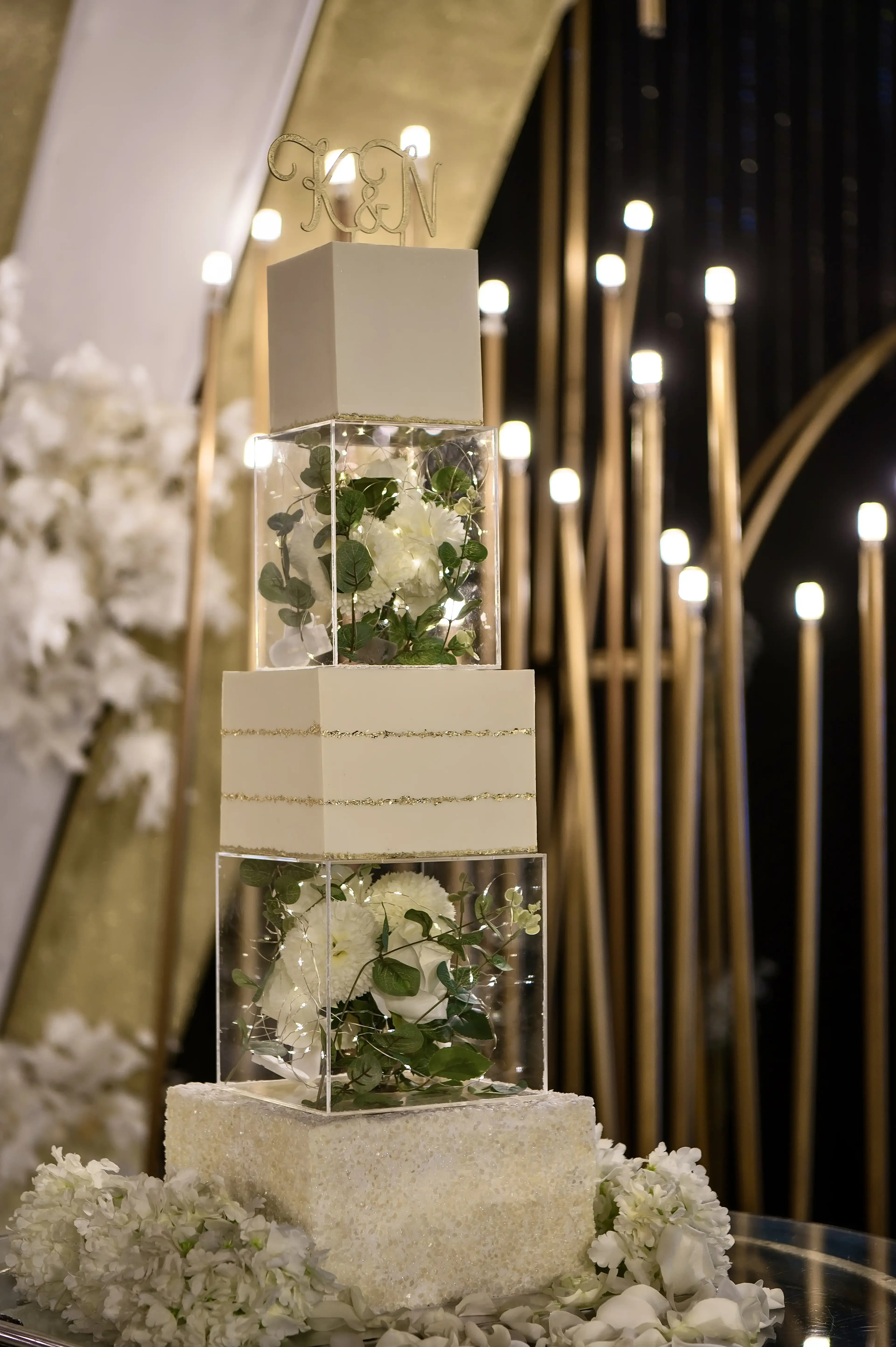 White and gold acrylic-cube tiered wedding cake with fresh white roses, greenery and fairy lights in the cube tiers, and a gold monogram topper