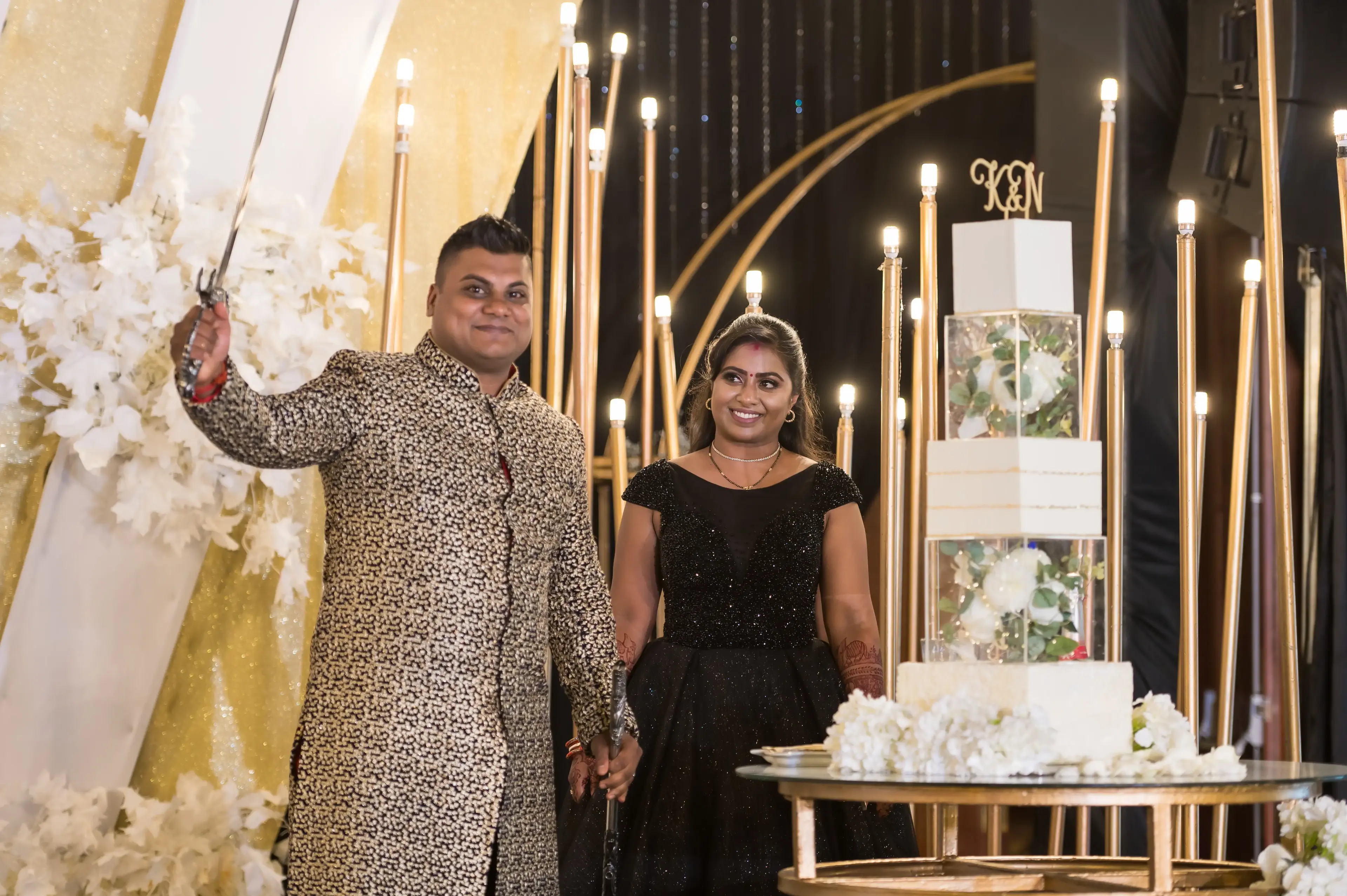 Bride and groom on stage, groom raising a ceremonial sword, beside a white and gold acrylic-cube tiered wedding cake with fresh white roses