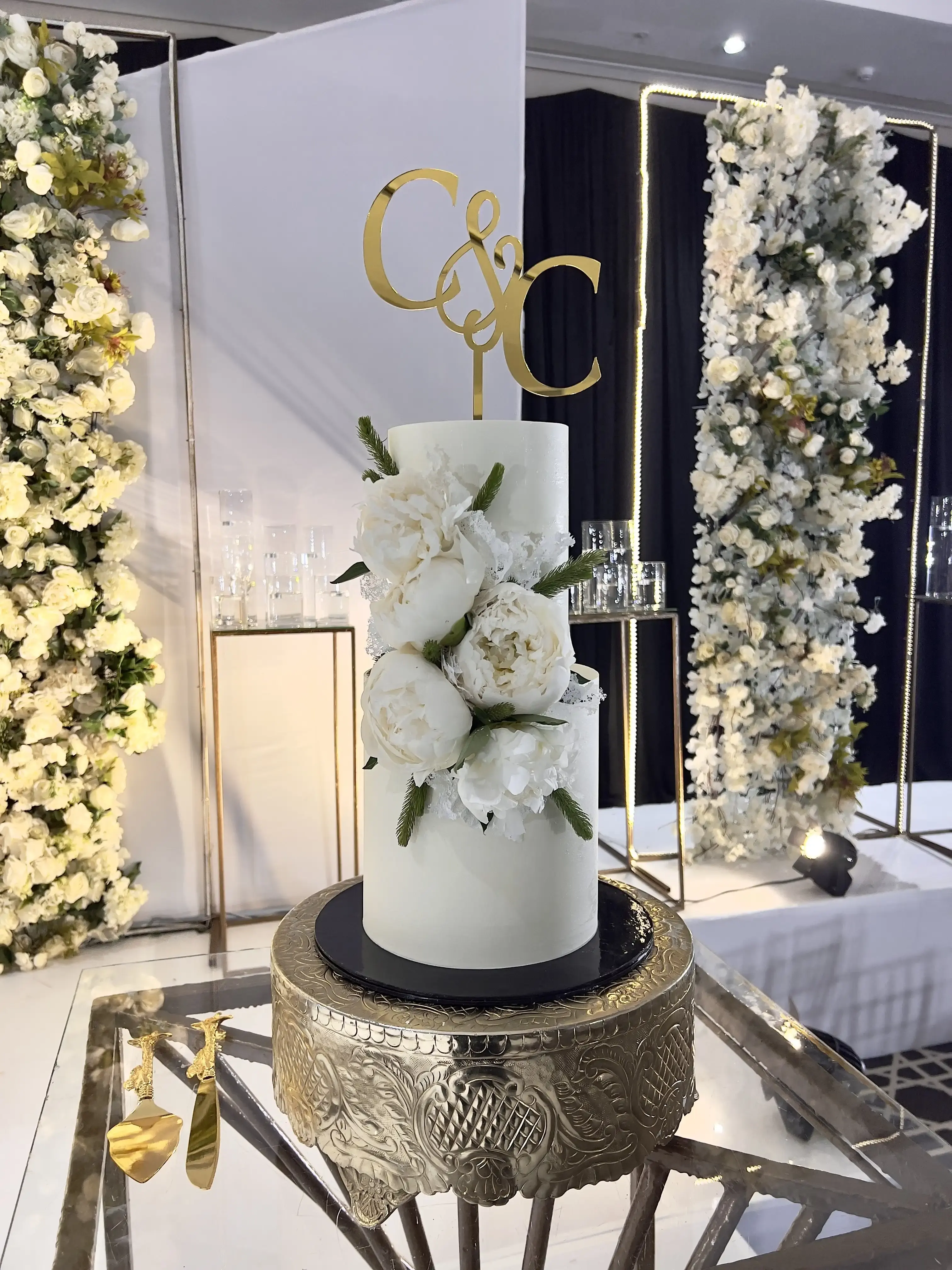 White wedding cake with fresh peonies and a gold script monogram topper against a floral wall