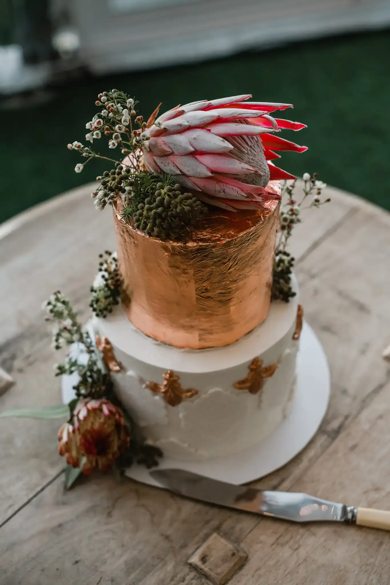 Two-tier wedding cake with a copper-foil top tier and fresh king protea, rustic outdoor table setting
