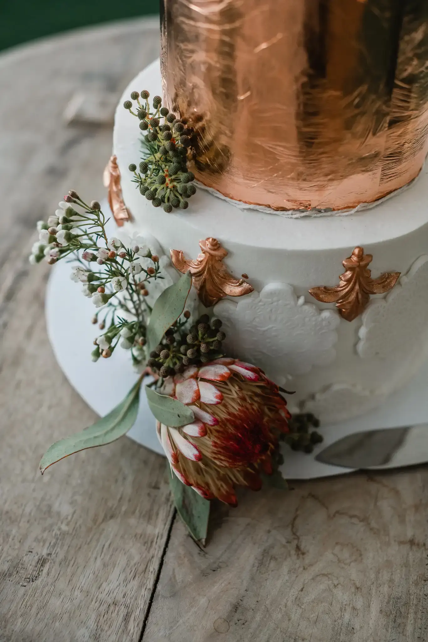 Detail of a copper-foil protea wedding cake showing gold fleur-de-lis piping and fresh florals