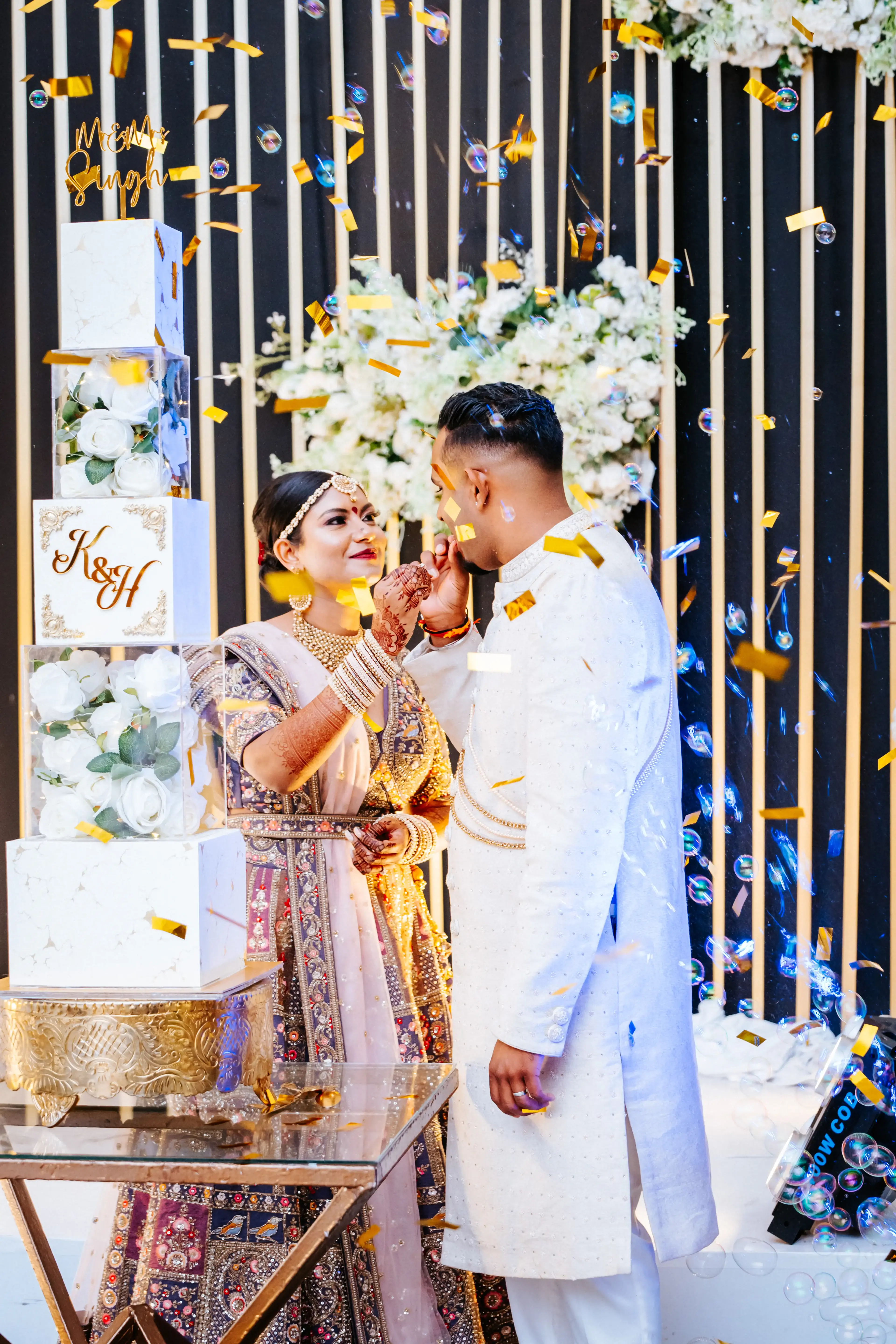 Bride and groom feeding each other cake beside an acrylic-cube floral wedding cake, confetti falling