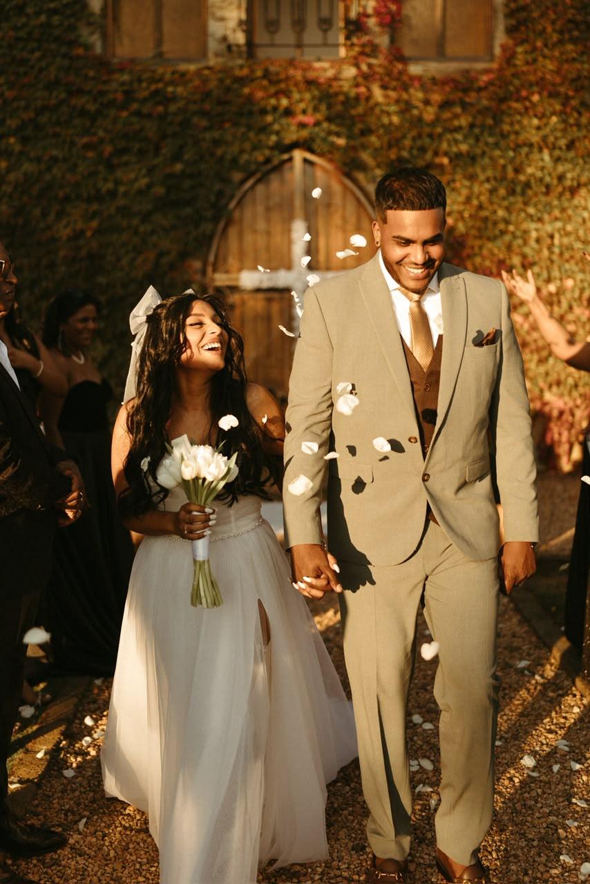 A couple laughing arm in arm as confetti falls, walking away from a stone chapel entrance