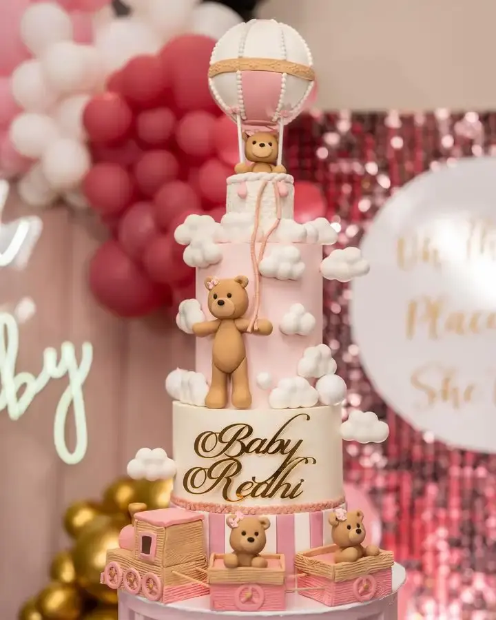 Pink four-tier baby shower cake with teddy bear figures, hot air balloon topper and piped clouds