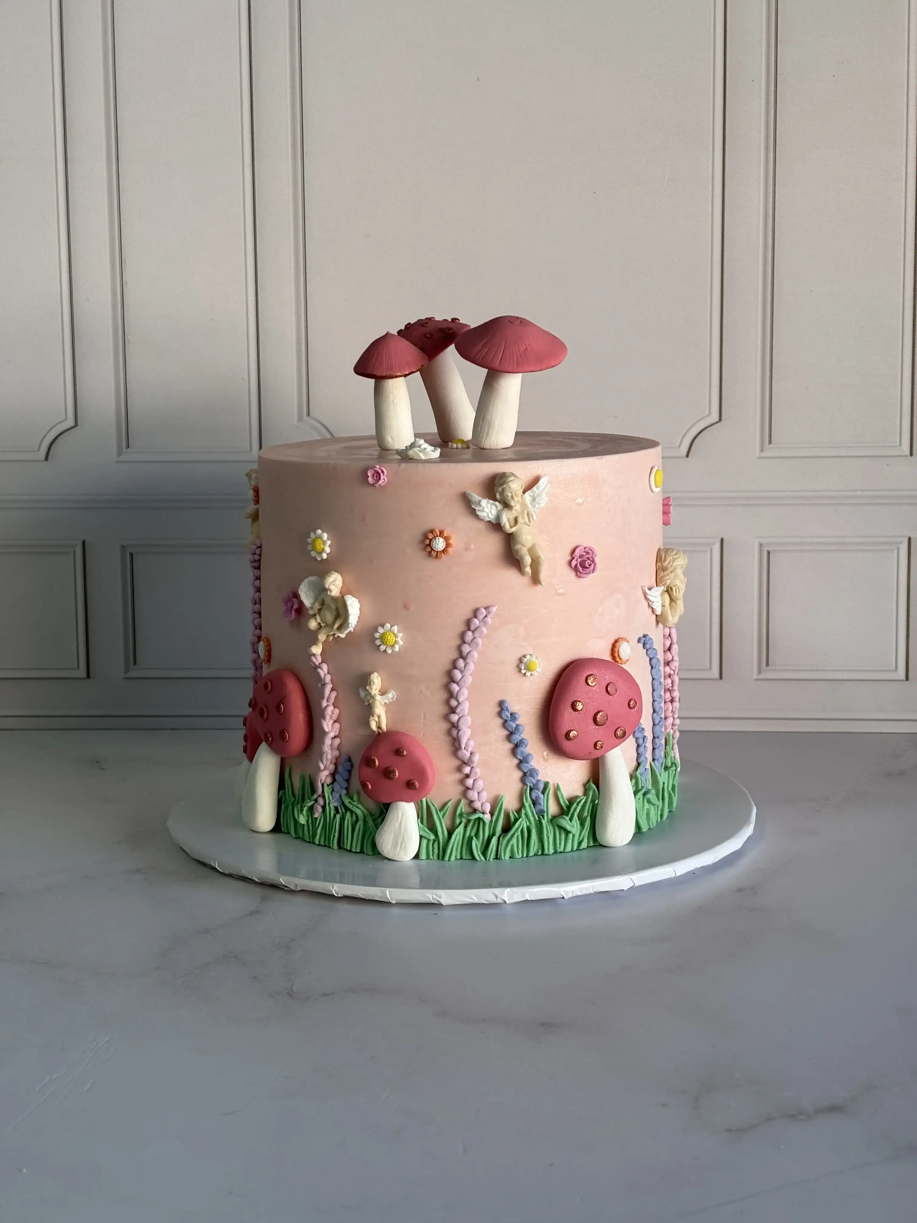 Pink cake decorated with fondant toadstools, cherub figures and piped flowers in a fairy-garden style